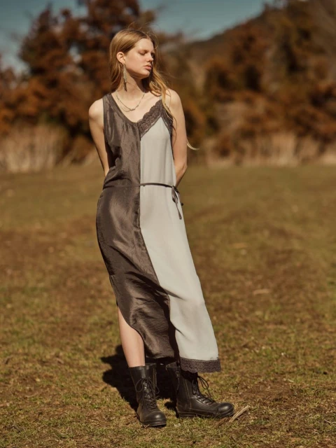 A young, blonde woman stands in a field wearing a two-tone black and light gray dress and black boots, with trees in the background.