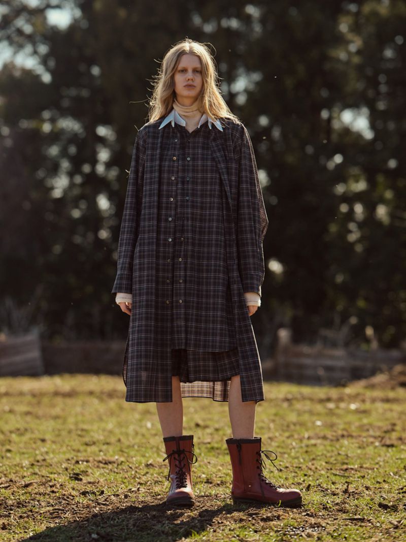 A blonde woman stands on a grassy field, wearing a plaid shirt-dress and lace-up boots, posing in an outdoor fashion shot.