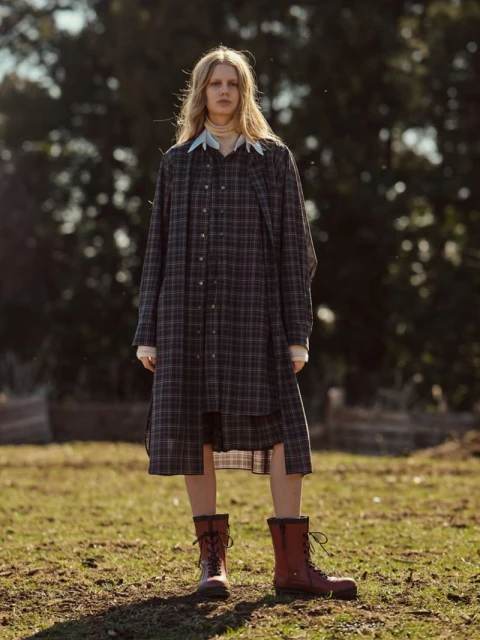 A blonde woman stands on a grassy field, wearing a plaid shirt-dress and lace-up boots, posing in an outdoor fashion shot.