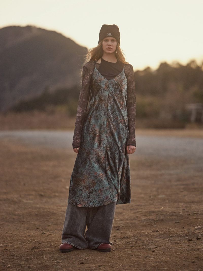 Model wearing a beanie, patterned slip dress over long sleeves, and wide-leg jeans in an outdoor landscape.