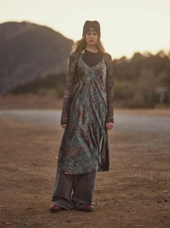 Model wearing a beanie, patterned slip dress over long sleeves, and wide-leg jeans in an outdoor landscape.