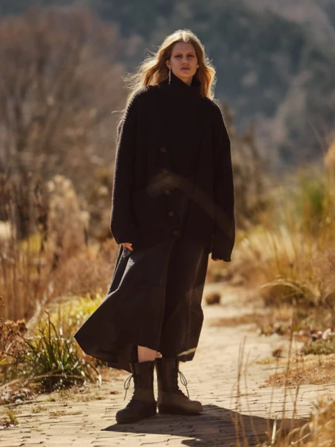 A full-length portrait of an adult woman posing on a stone path. She is wearing a long, dark cardigan and dress with lace-up boots.