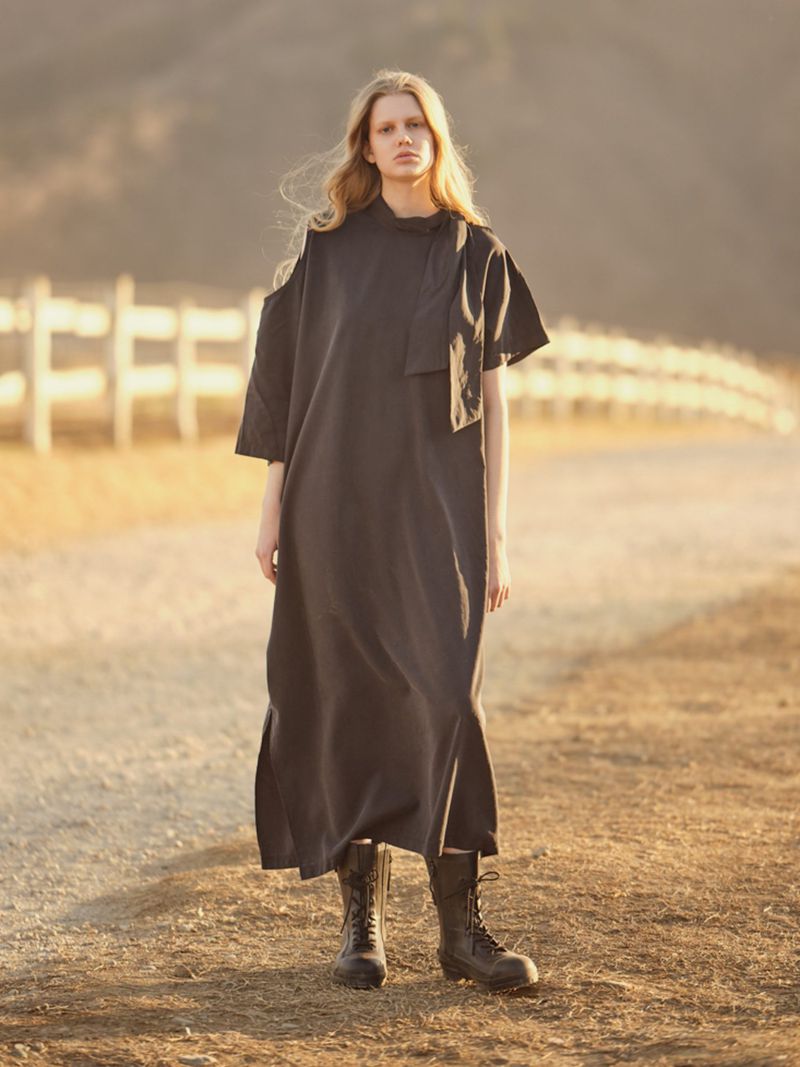 A full-length portrait captures a young adult female model in a black dress and boots on a dirt road with a rural backdrop.