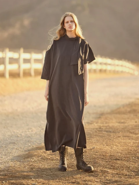 A full-length portrait captures a young adult female model in a black dress and boots on a dirt road with a rural backdrop.
