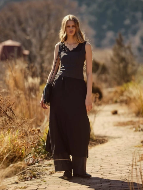 Woman in black top and skirt walks on brick path in sunny outdoor fashion shoot with natural lighting and a blurred background.
