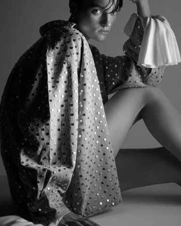Black and white portrait of a person with short dark hair wearing a polka dot jacket with puffy sleeves sitting indoors.