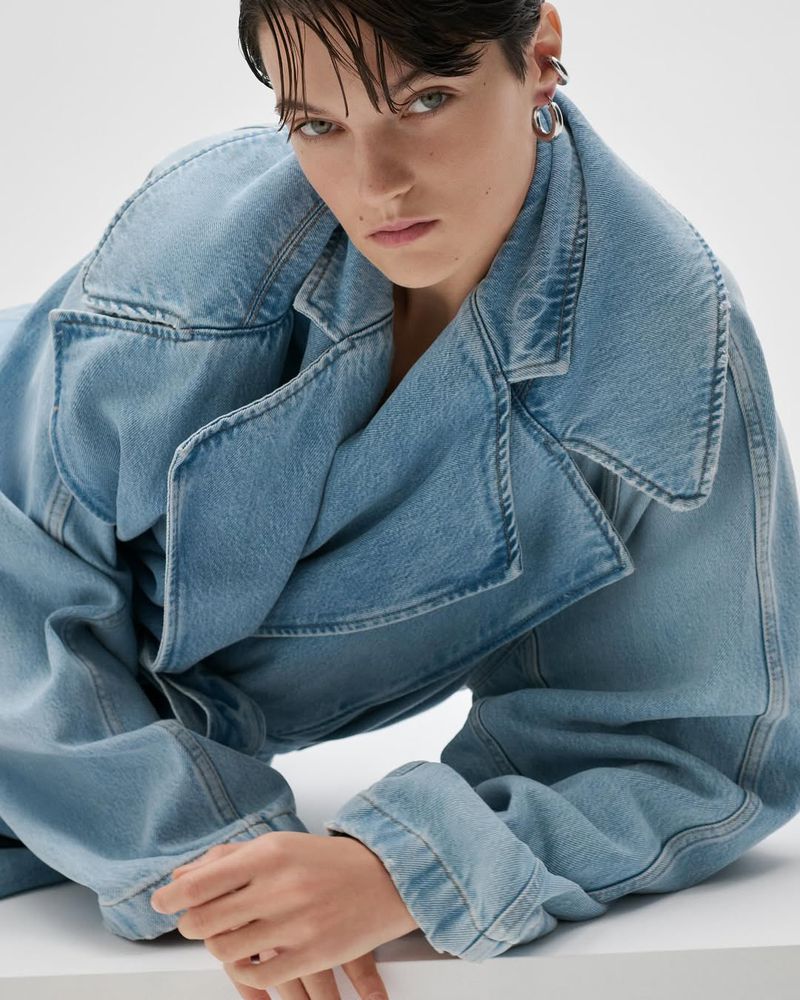 Woman poses wearing a light blue denim trench coat indoors against a white backdrop, showcasing a fashion-forward and casual style.