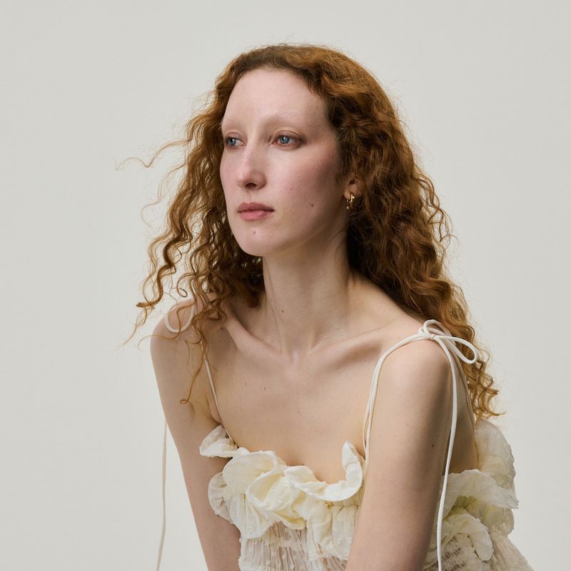 Portrait of a red-haired woman with pale skin wearing a frilly, textured light yellow top in a photo studio setting.