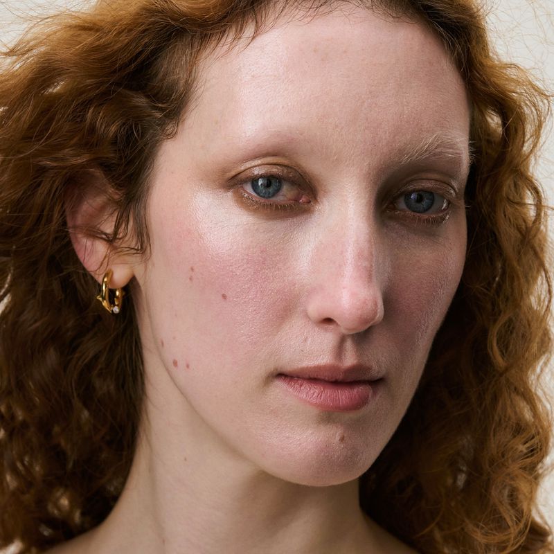 Close-up portrait of a woman with curly red hair and blue eyes wearing gold hoop earrings against a neutral background