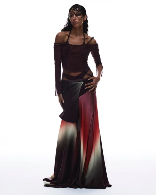 Full-length studio shot of a woman with dark hair wearing a dark brown, long-sleeved top and a long, gradient skirt in a fashion pose.