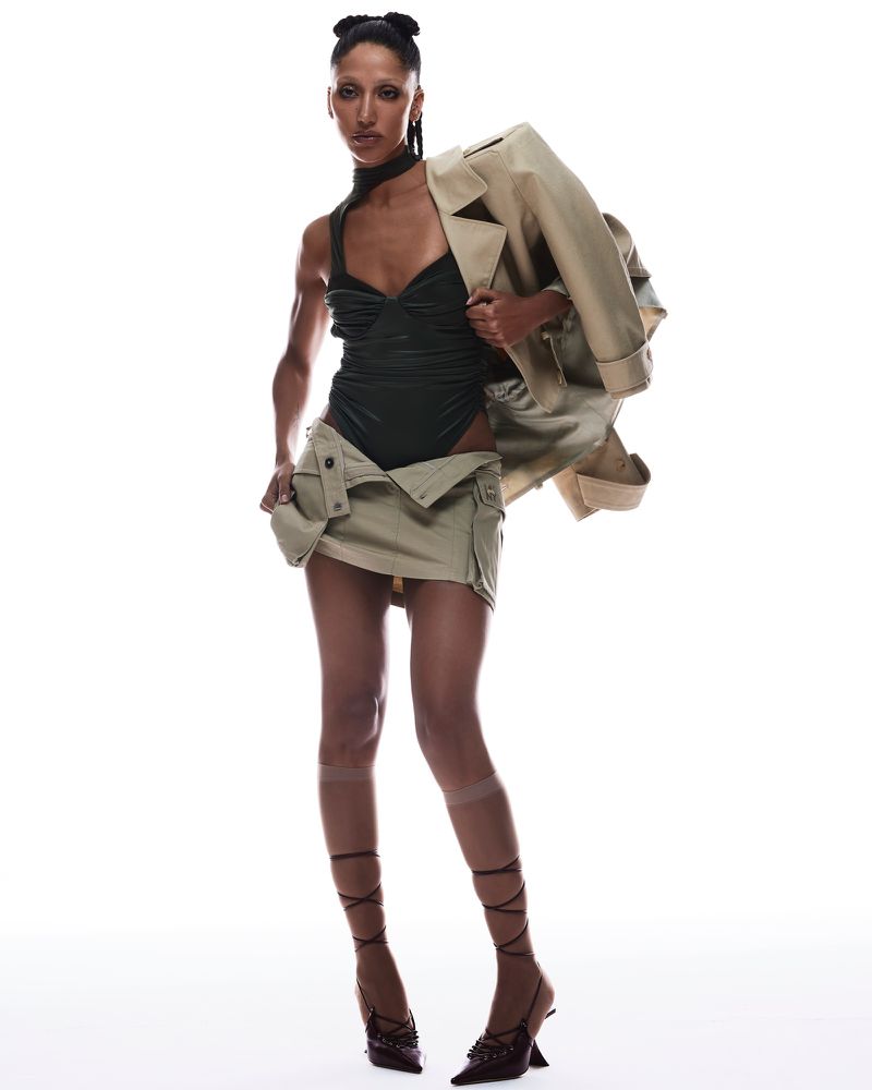 Woman in black bodysuit, tan mini skirt, and jacket poses confidently in a fashion shot against a bright white background.