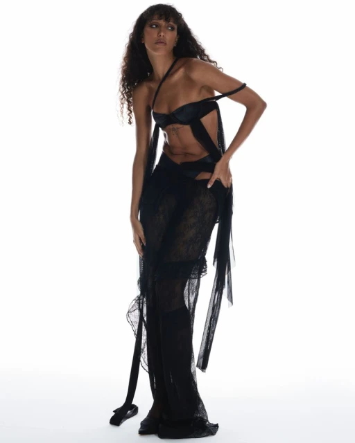 Woman in a black sheer dress poses in a photo studio against a white backdrop, showcasing a fashion-forward and elegant look.