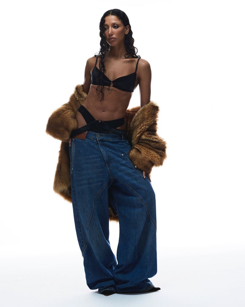 Fashion editorial shot of a woman in black lingerie, an open brown fur coat, and oversized blue jeans against a bright white backdrop.