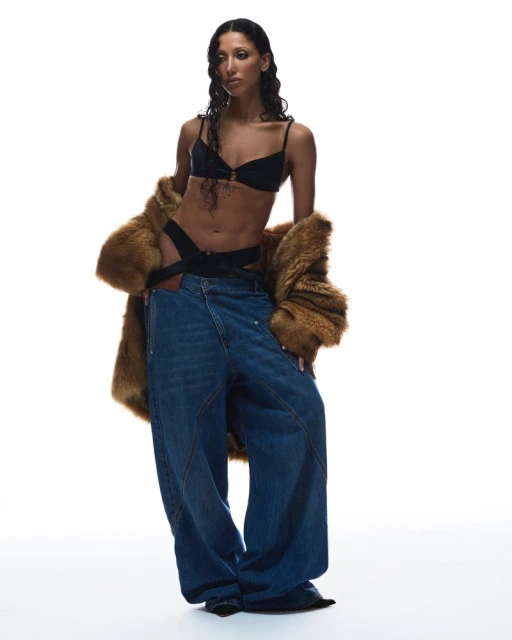 Fashion editorial shot of a woman in black lingerie, an open brown fur coat, and oversized blue jeans against a bright white backdrop.