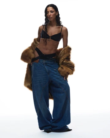 Fashion editorial shot of a woman in black lingerie, an open brown fur coat, and oversized blue jeans against a bright white backdrop.
