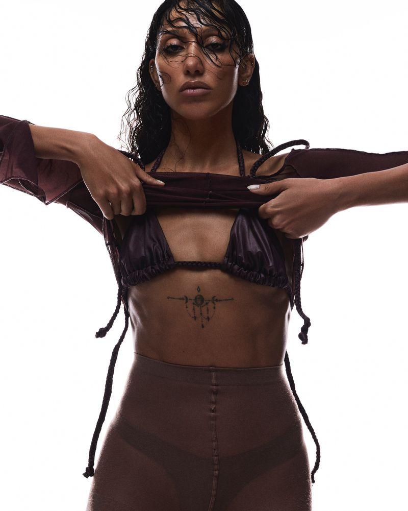 A woman with wet dark hair is wearing a dark bra top, bottoms, and a loose long-sleeve top while posing against a white backdrop.