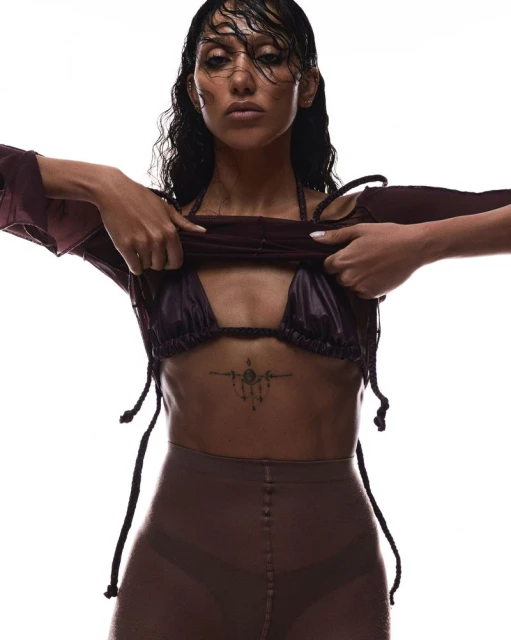 A woman with wet dark hair is wearing a dark bra top, bottoms, and a loose long-sleeve top while posing against a white backdrop.