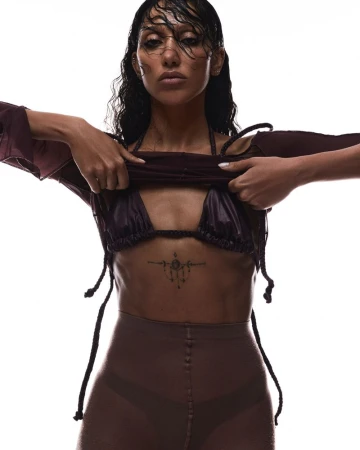 A woman with wet dark hair is wearing a dark bra top, bottoms, and a loose long-sleeve top while posing against a white backdrop.