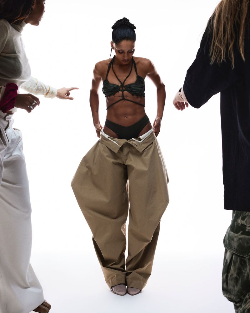 Fashion shot of a woman in a bikini bottom and a person holding up oversized pants in front of her, flanked by two other figures.