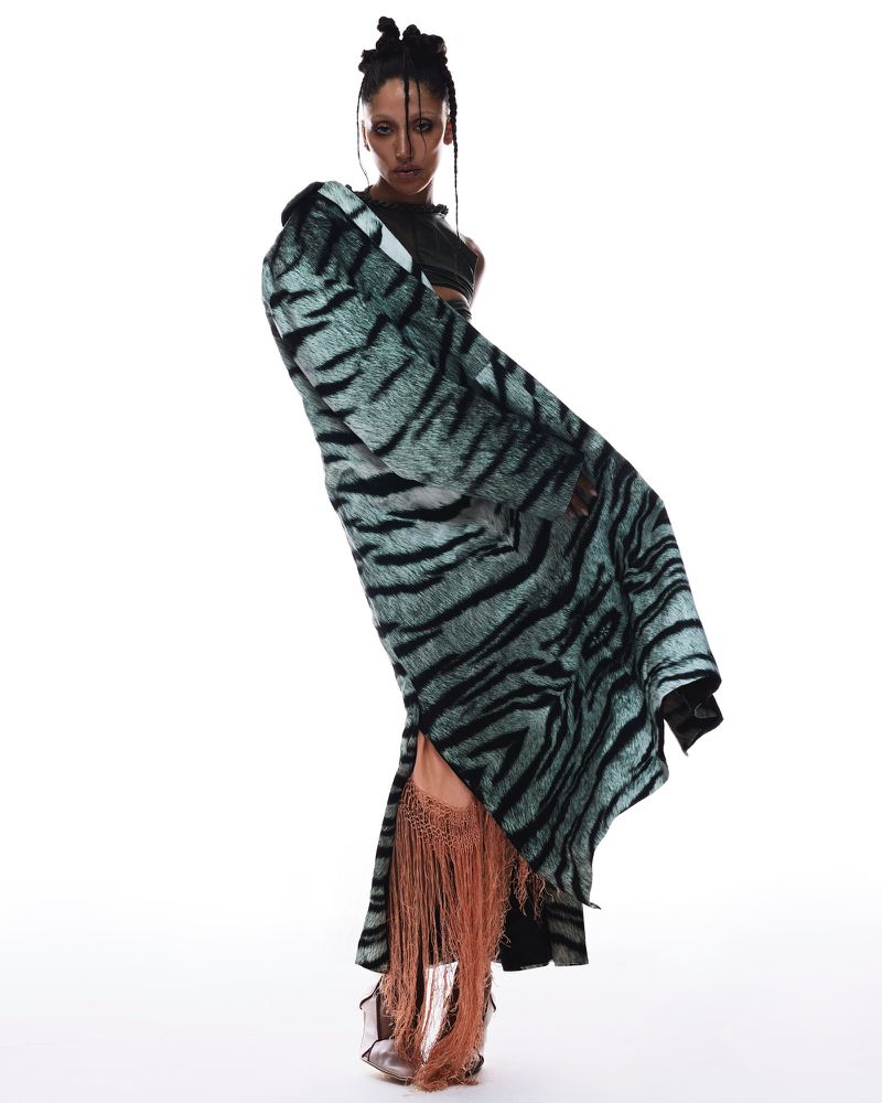 Model wearing a teal patterned coat and fringe skirt standing against a white studio background