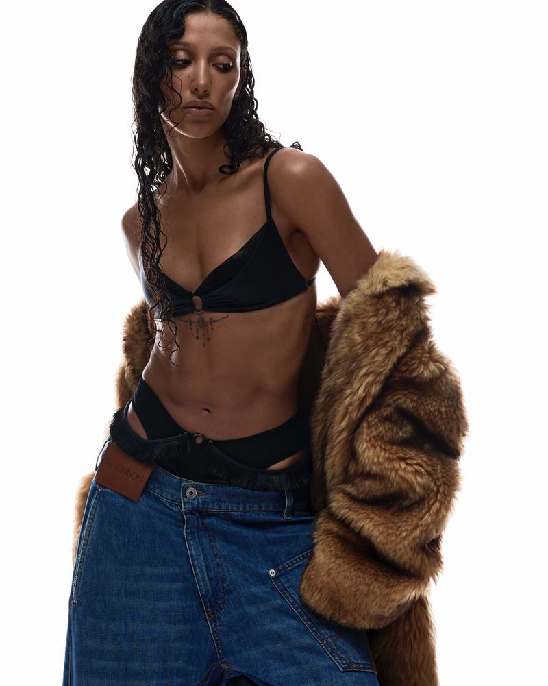 A woman with wet dark hair poses in a black bikini top and oversized denim jeans with a fur coat against a bright white backdrop.