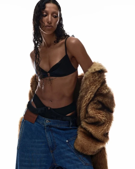 A woman with wet dark hair poses in a black bikini top and oversized denim jeans with a fur coat against a bright white backdrop.