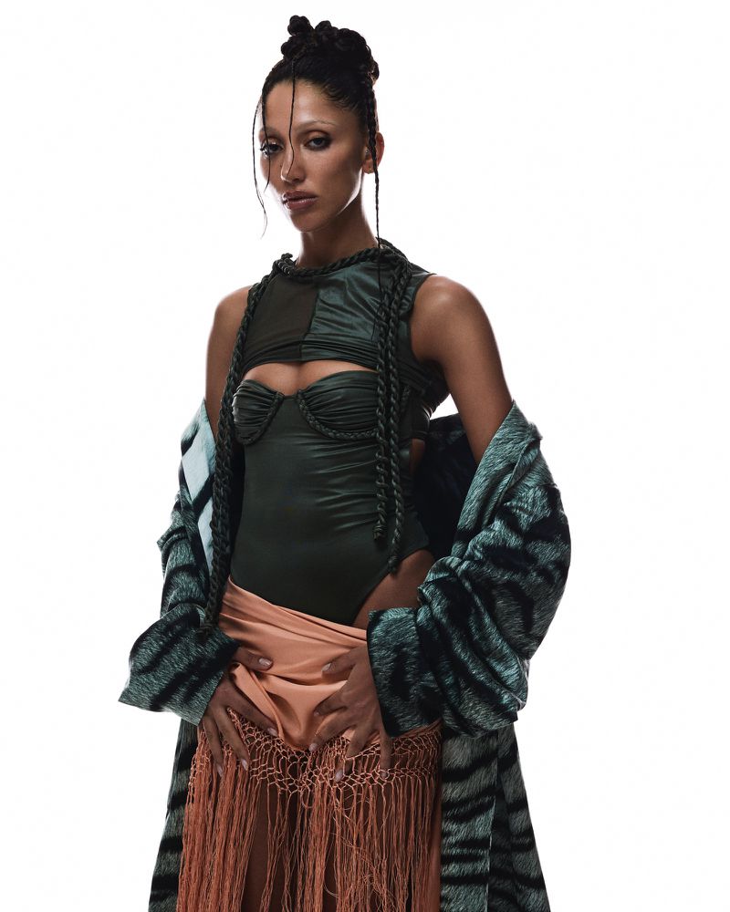 Fashion shot of a slender adult woman with an elaborate hairstyle wearing a green bodysuit and coat against a white backdrop.