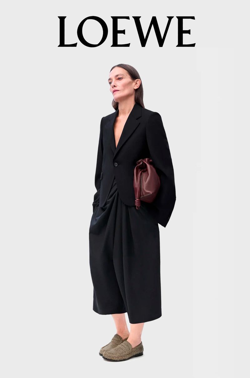 Brunette female model wears a black suit with wide black pants, holding a maroon bag, standing in a photo studio for LOEWE advertisement.