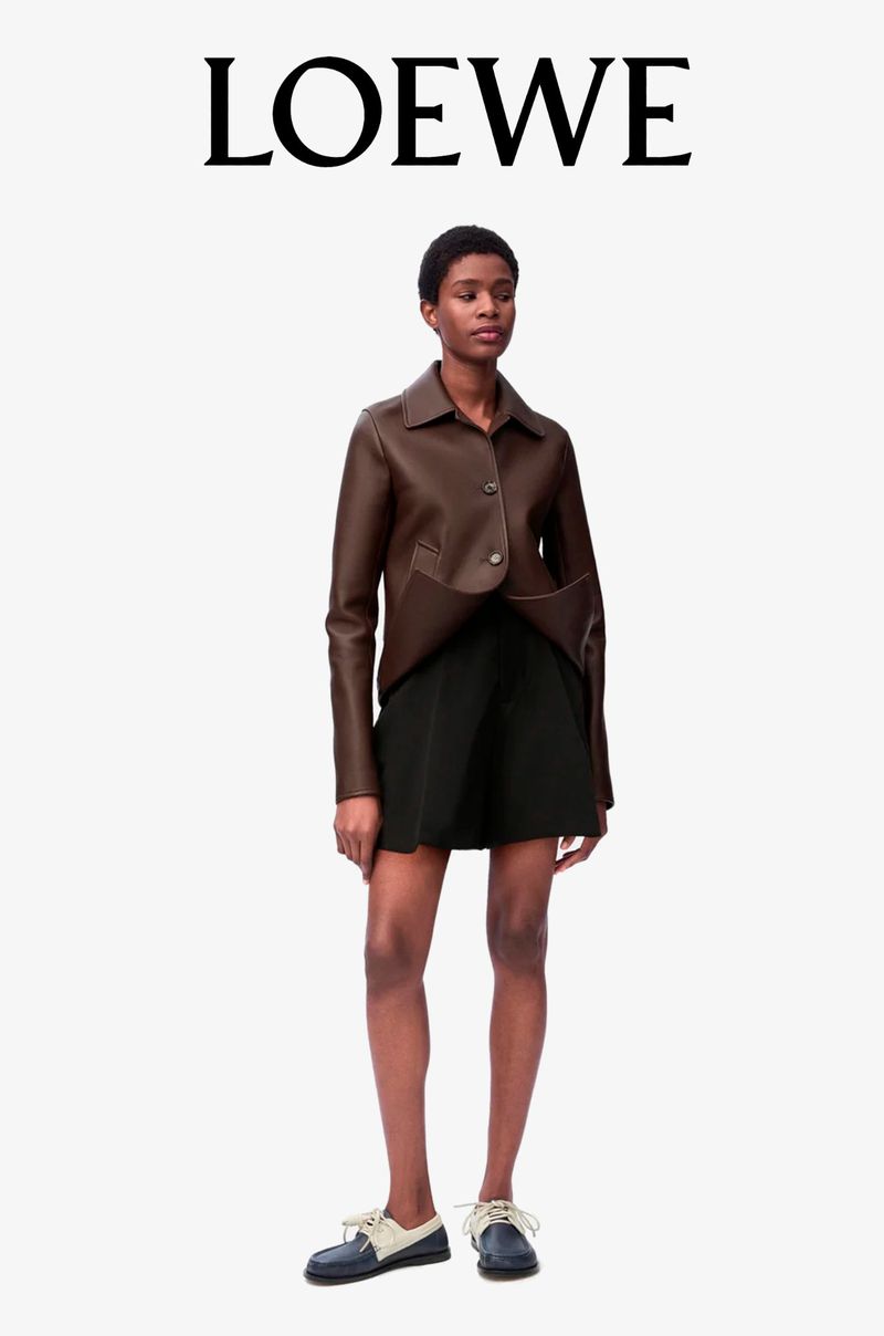 A woman stands in a studio, wearing a brown jacket, black shorts, and loafers, under the brand name LOEWE in a fashion editorial shot.