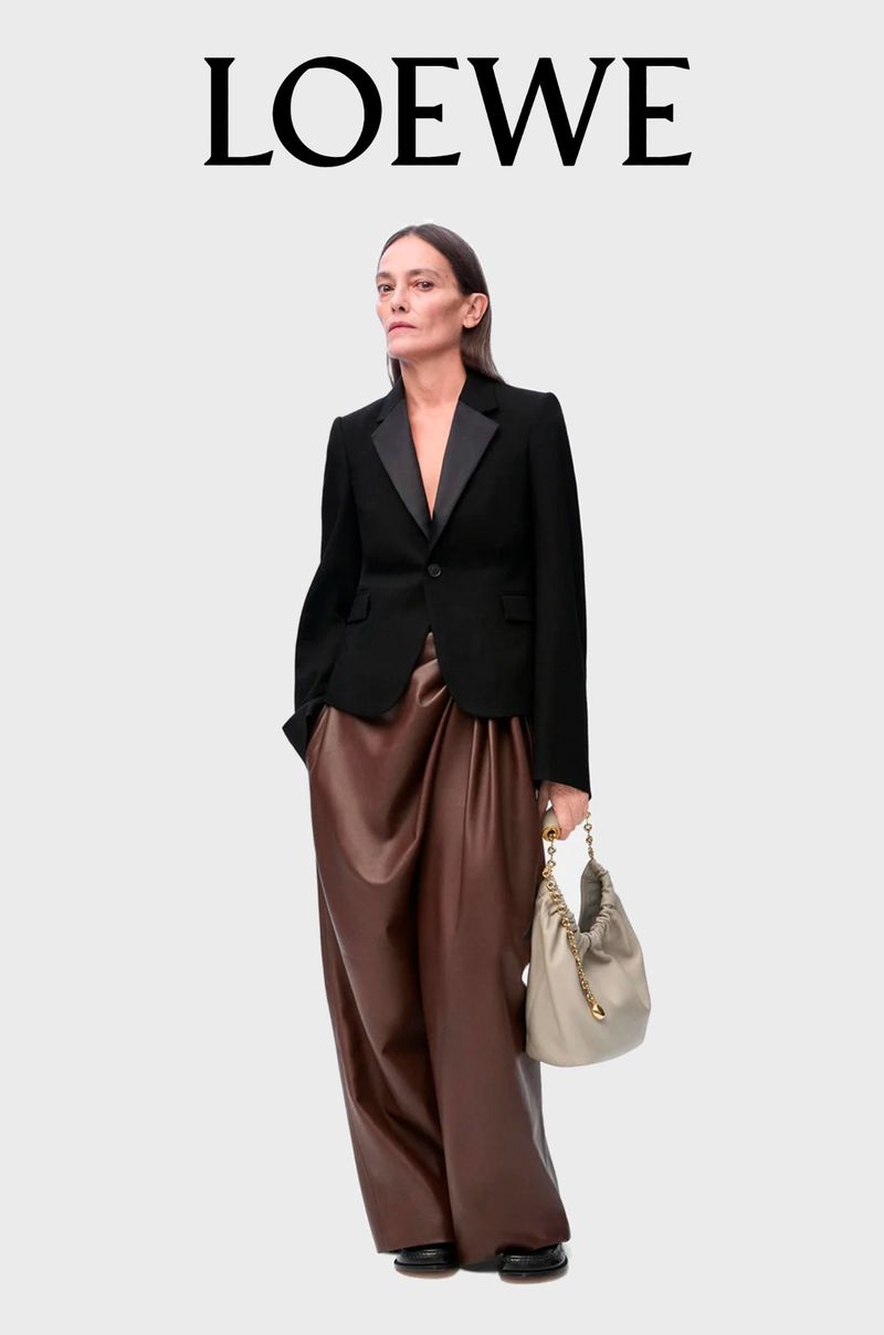 A woman poses in a black blazer, brown pants, and a beige bag under the LOEWE logo. She has straight brown hair and pale skin.