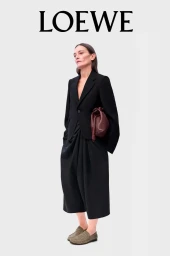 Model wearing a black blazer and gathered skirt holding a brown clutch against a plain white background with Loewe typography above