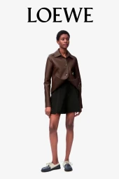 Model wearing a brown leather jacket and black shorts standing against a plain white studio background.