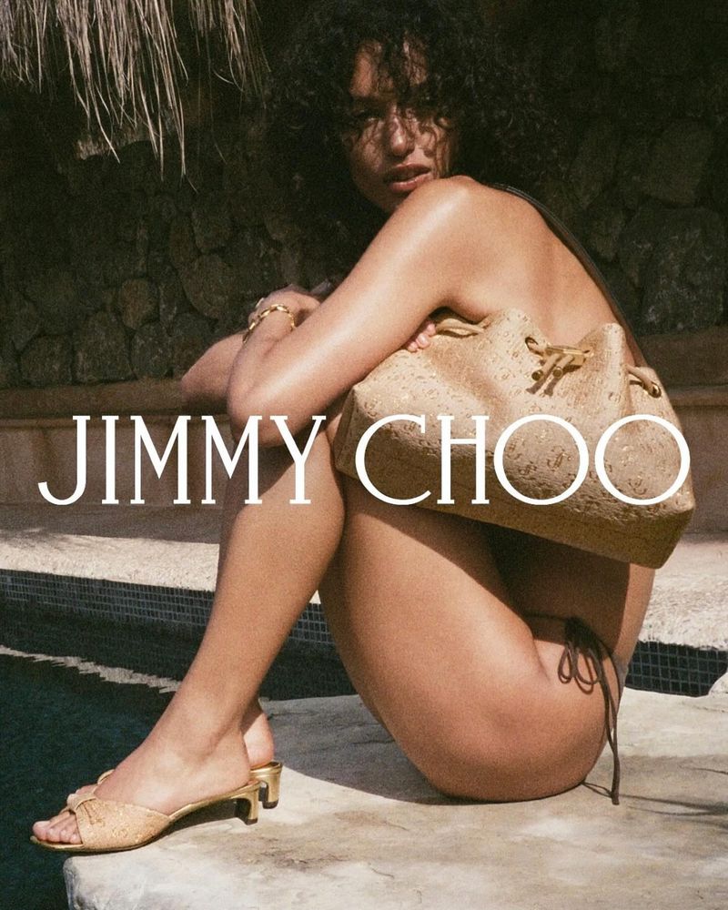 Woman poses with a Jimmy Choo bucket bag and gold heels on a tiled surface near a pool, set against a textured stone wall backdrop.
