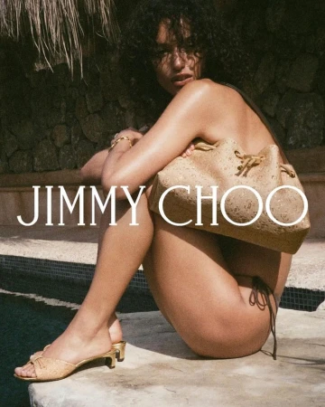 Woman poses with a Jimmy Choo bucket bag and gold heels on a tiled surface near a pool, set against a textured stone wall backdrop.