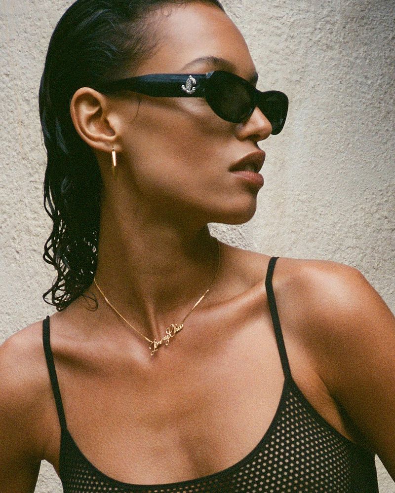 Woman in a black mesh top wearing sunglasses and a gold necklace stands in front of a neutral wall in this close-up studio shot.