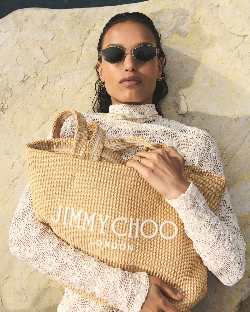 A model with sunglasses and a white lace top poses with a Jimmy Choo woven tote bag on a rock surface outdoors.