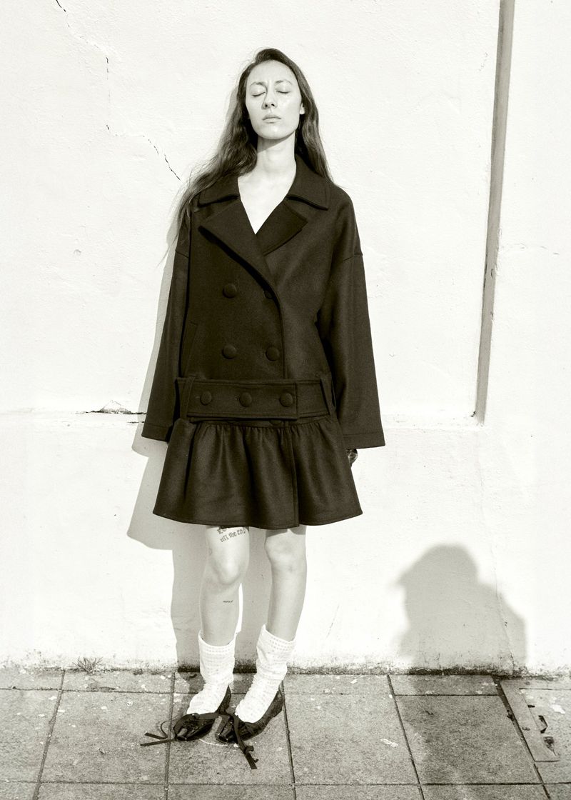 Woman in a black coat and skirt poses in front of a textured white wall, wearing white socks and black shoes with closed eyes.