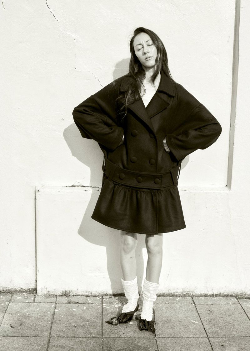 Monochrome full shot of woman in dark coat with skirt, white socks, and dark shoes posing against a white weathered wall on a concrete sidewalk.