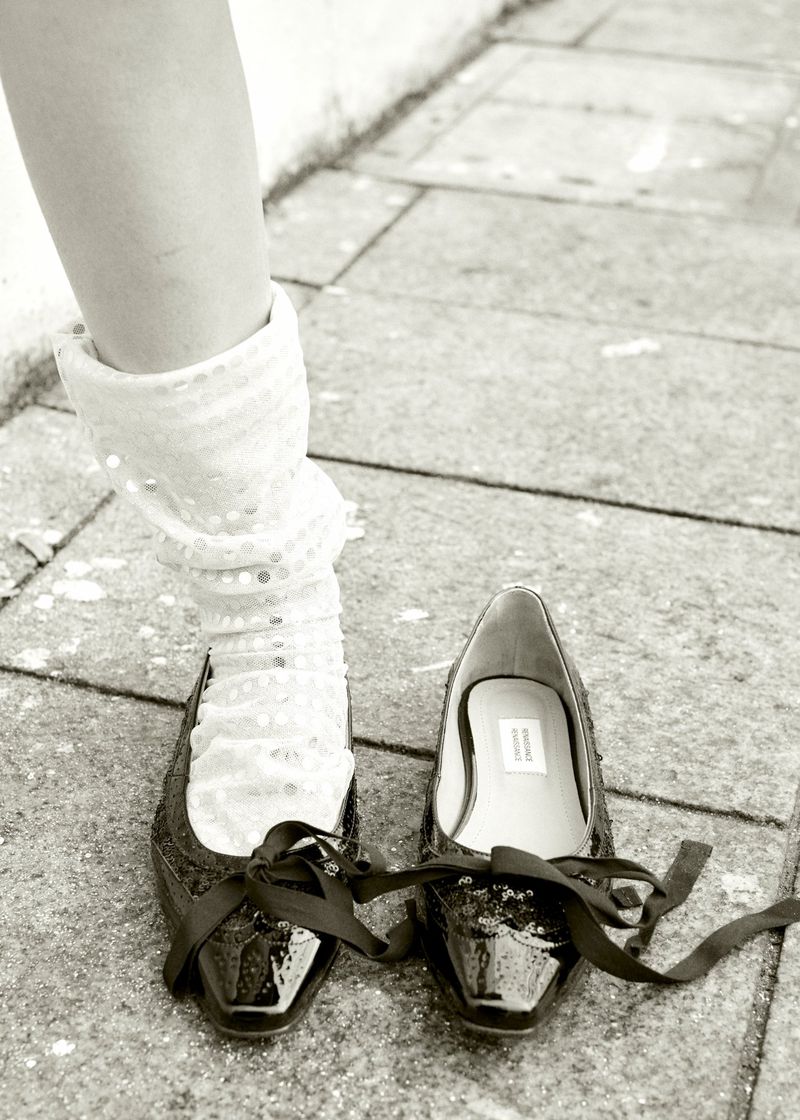 Eye-level shot of a person's lower leg in a sparkly sock standing next to a pair of patent leather ballet flats with ribbon ties on a stone surface.