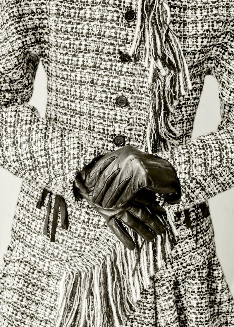 Close-up studio shot of a tweed skirt suit with button closure, fringe, and black leather gloves, creating a chic fashion ensemble.
