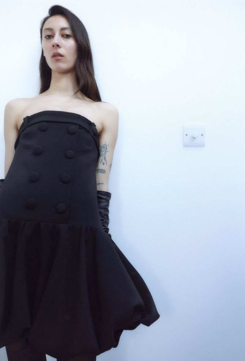 Woman poses in black strapless dress with gloves against white wall, fashion shoot, neutral expression, indoor lighting, studio.