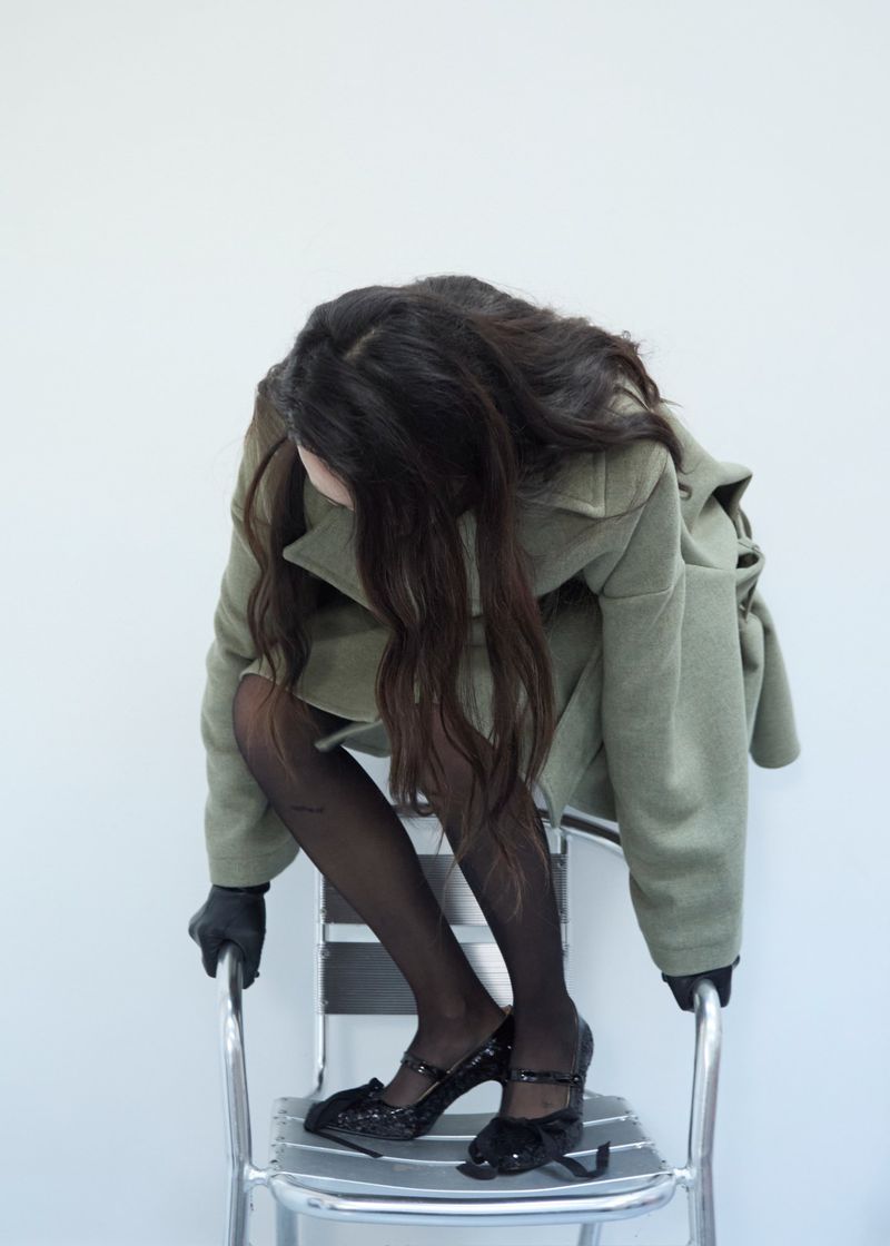 Fashionable woman in an olive overcoat, black stockings, and patent-leather heels sits on a chrome chair against a white background.