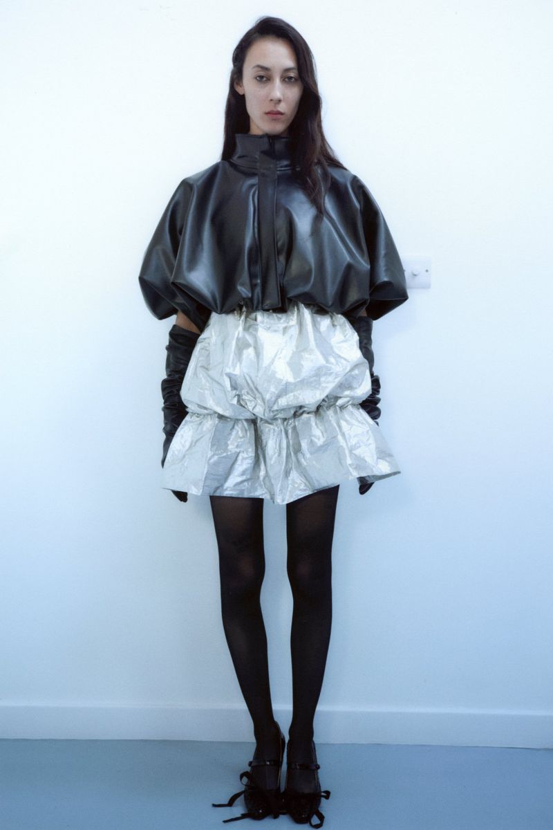Young woman in a black leather top and silver skirt poses against a white wall, showcasing a fashion-forward, contemporary style.