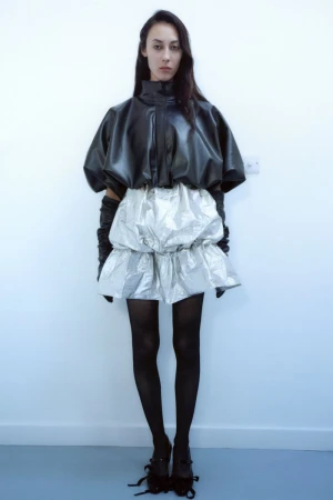 Young woman in a black leather top and silver skirt poses against a white wall, showcasing a fashion-forward, contemporary style.