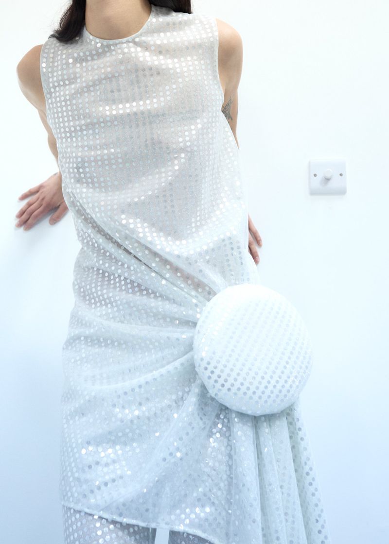 Adult woman wearing a formal white sequined sleeveless dress with a large decorative circular accent posing indoors against a white wall.