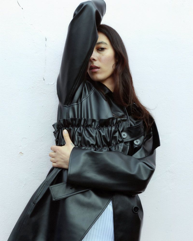 A woman in a black faux leather coat poses with her arm raised against a textured light pink wall in an indoor setting.