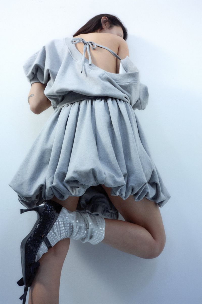 Woman in a gray puffball dress and heeled shoes poses against a white wall, showcasing a contemporary fashion style with a unique silhouette.