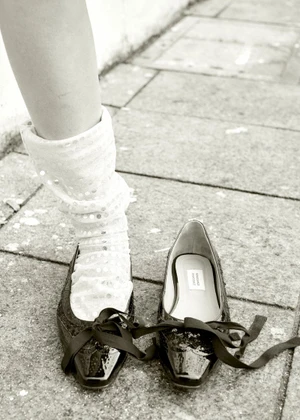 Close-up of a leg in a white sequined slouch sock with a black patent leather flat and matching shoe on stone pavement.