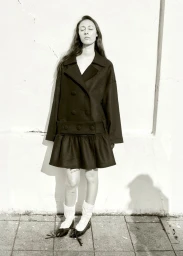 Model in a black double-breasted coat standing against a light wall in full-length view.