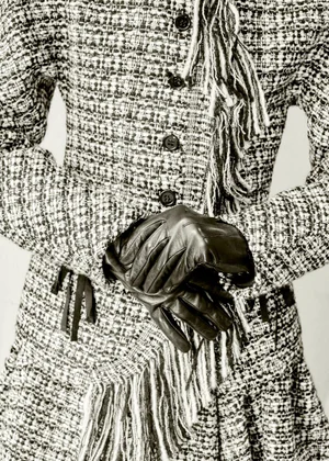 Close-up of a person wearing a black and white tweed jacket and black leather gloves against a plain background